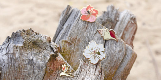 flower and shell enamel pins sitting on a piece of driftwood on the beach